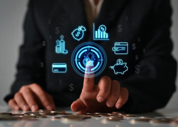 A person in a suit touches a digital financial icon on a virtual screen, surrounded by symbols of money, charts, payment, and savings, with coins scattered on the table in the foreground. | GRAMHIR