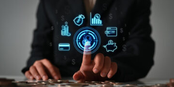 A person in a suit touches a digital financial icon on a virtual screen, surrounded by symbols of money, charts, payment, and savings, with coins scattered on the table in the foreground. | GRAMHIR
