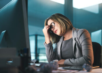 A woman in business attire sits at a desk in front of a computer, looking stressed with her hand on her forehead. Crumpled papers are scattered on the desk, and office windows are in the background. | GRAMHIR