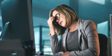 A woman in business attire sits at a desk in front of a computer, looking stressed with her hand on her forehead. Crumpled papers are scattered on the desk, and office windows are in the background. | GRAMHIR