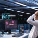 A person in an office holds their head in distress while looking at computer screens displaying red warning messages and text, including "System Hacked," indicating a cybersecurity breach. | GRAMHIR