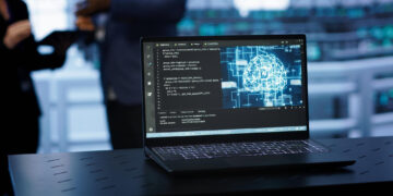 A laptop on a desk displays code and a digital brain graphic. In the blurred background, two people stand and interact in a modern, high-tech setting. | GRAMHIR
