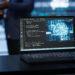 A laptop on a desk displays code and a digital brain graphic. In the blurred background, two people stand and interact in a modern, high-tech setting. | GRAMHIR
