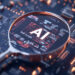 A magnifying glass hovers over a computer circuit board, highlighting the glowing letters “AI” to symbolize artificial intelligence technology and digital innovation. | GRAMHIR